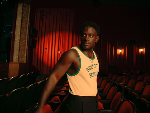 A person in a tank top with "Soul of the Seventies" stands in an empty theater with red seats and curtains, lit by warm lighting.