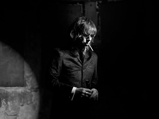 A person in a dark suit stands in partial spotlight, smoking, in a dimly lit room. Black and white mood evokes a sense of mystery.