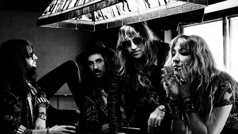 Four people with long hair and sunglasses gather around a table under a stained glass lamp in a dimly lit room, exuding a relaxed vibe.