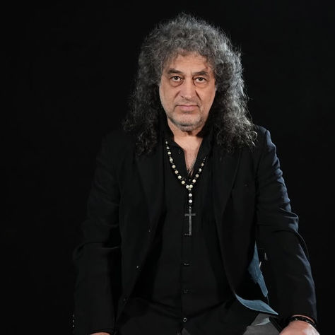 Gray-haired man in a black suit and cross necklace poses against a dark background with a serious expression