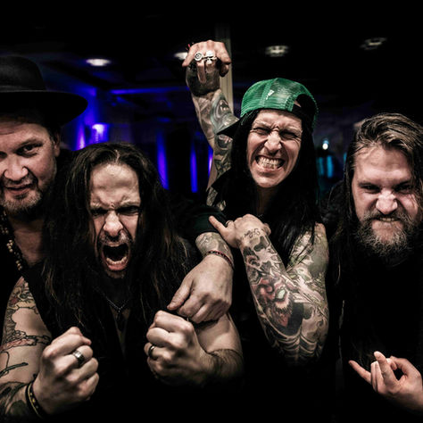 Four tattooed men pose fiercely in a dark club, one in a green cap and one in a black hat, fists raised under blue lights.