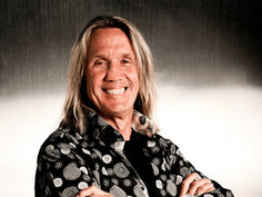 Smiling man with long hair in patterned shirt, arms crossed. Gray gradient background.