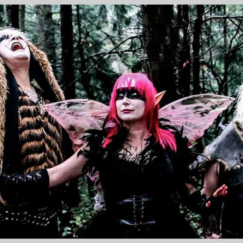 Three people in fantasy costumes with dramatic expressions in a forest. One has red wings, another wears fur, all in dark outfits.