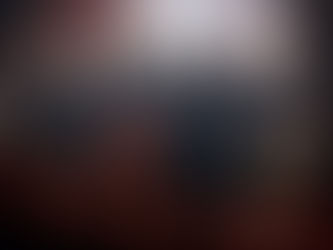 Seven people stand arm-in-arm on stage, smiling. They wear dark shirts, and one has red pants. Instruments and a dim background are visible.