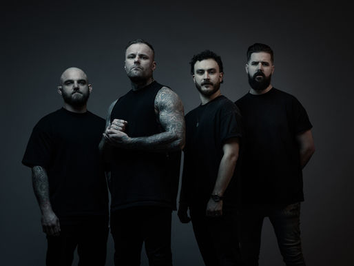 Four serious men in black T-shirts pose in a dark studio, one with tattooed arms and clasped hands.