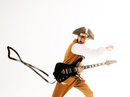 Man in period costume energetically plays electric guitar against a bright white background, expressing excitement and intensity.