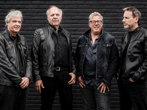 Four men in black leather jackets stand against a dark brick wall, posing confidently. The mood is serious and focused.