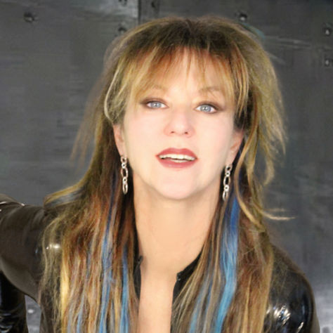 Person with multicolored hair and bright earrings in a shiny black jacket, smiling against a textured gray background.