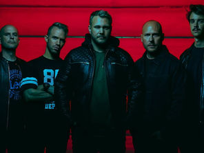 Five men in dark jackets stand against a red background, looking serious. One wears a shirt with "89" text. The mood is intense.