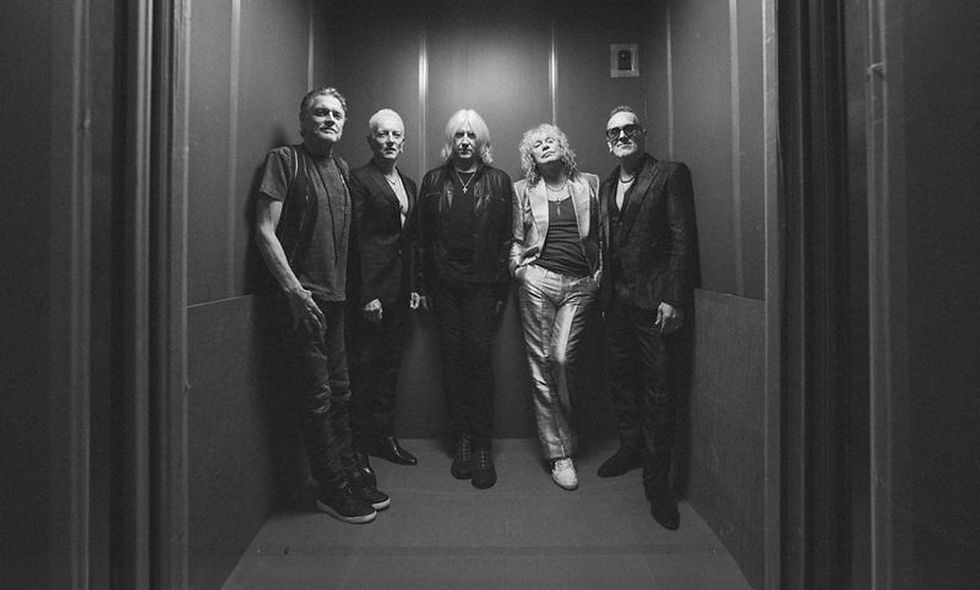 Five people stand confidently in an elevator, wearing dark clothing. The black-and-white image has a cool, relaxed mood.