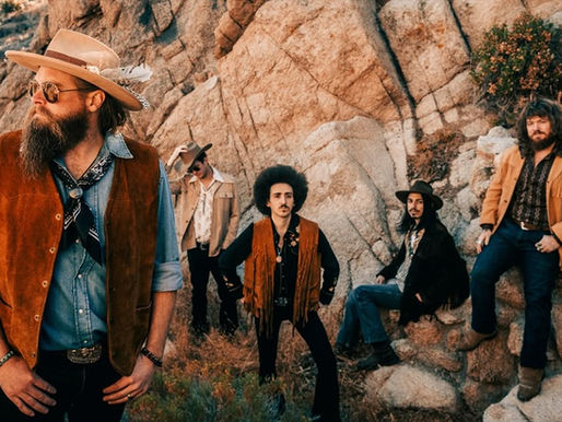 Five men in cowboy attire stand and sit on rocky terrain, wearing hats and fringe vests. The scene exudes a vintage Western vibe.