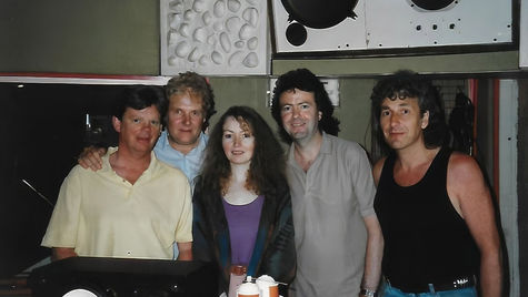 Five people smiling in a recording studio with speakers, wearing casual clothes. The atmosphere is relaxed and friendly.