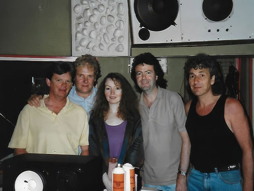 Five people smiling in a recording studio with speakers, wearing casual clothes. The atmosphere is relaxed and friendly.