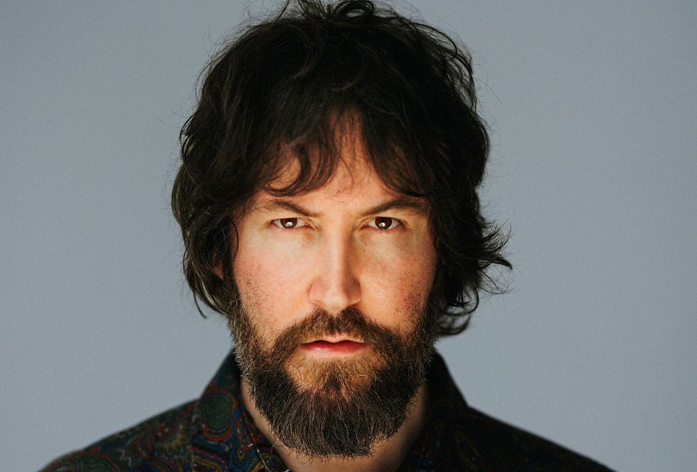 Close-up portrait of a bearded man with shaggy dark hair and an intense stare against a plain gray background