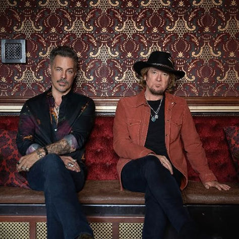 Two men sit on a red velvet bench against ornate wallpaper. One wears a black shirt, the other a hat and orange jacket, conveying a relaxed mood.
