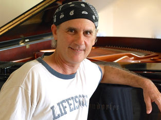 Man in a white "LIFE IS GOOD" shirt and bandana smiles, leaning on a grand piano. Warm, indoor setting with soft lighting.