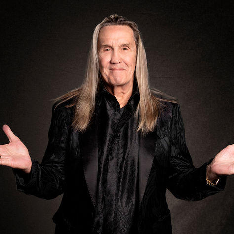 Older man with long gray hair in a black suit, arms outstretched in a dark studio, smiling slightly.