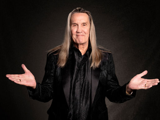 Older man with long gray hair in a black suit, arms outstretched in a dark studio, smiling slightly.