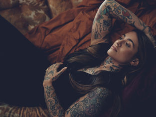 Tattooed woman reclines on orange bedding, hand on chest, looking thoughtful. Warm tones and floral patterns create a relaxed mood.