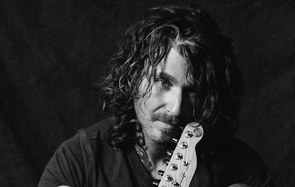 Man with curly hair leans on a guitar neck, looking thoughtful in a black and white photo. Dark background, moody atmosphere.