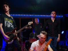 Four musicians pose with instruments in a dimly lit studio. One wears a "Love Kills" shirt. Background lights add a dramatic mood.