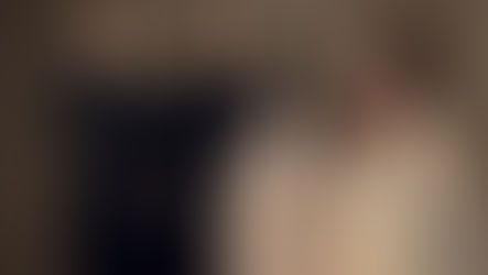 Four people in formal attire pose elegantly against a brown backdrop. The women wear ornate beige dresses, the men in black suits.