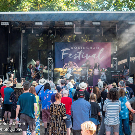 Crowd watches live band on an outdoor stage at Wokingham Festival, with smoke, trees, and a colorful banner.