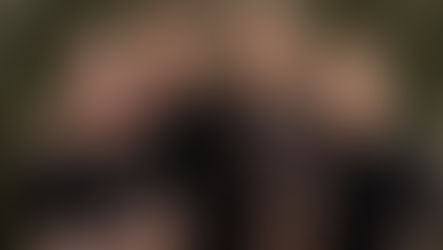 Four men with tattoos stand against a weathered green wall. Three wear black shirts, one wears brown. Serious expressions, casual vibe.