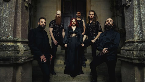 Six people in dark attire pose on stone steps of an old building. The setting is moody, with muted colors and an imposing vibe.