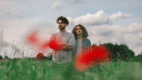 Two men stand in a green field with red flowers, looking thoughtful. The sky is cloudy, creating a calm and serene atmosphere.