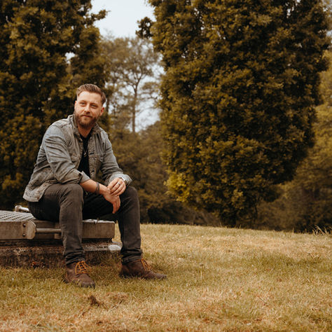 Man in denim jacket sits on a concrete bench in a grassy park, with tall trees behind him and a calm, thoughtful expression.