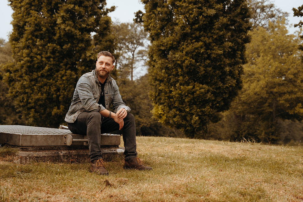 Man in denim jacket sits on a concrete bench in a grassy park, with tall trees behind him and a calm, thoughtful expression.
