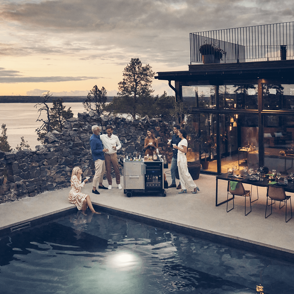 Terrasse avec piscine au crépuscule : un groupe d’amis se rassemble autour d’un bar mobile Dometic MoBar 550S pour préparer des cocktails, devant une maison d’architecte vitrée avec vue sur un lac et les collines.