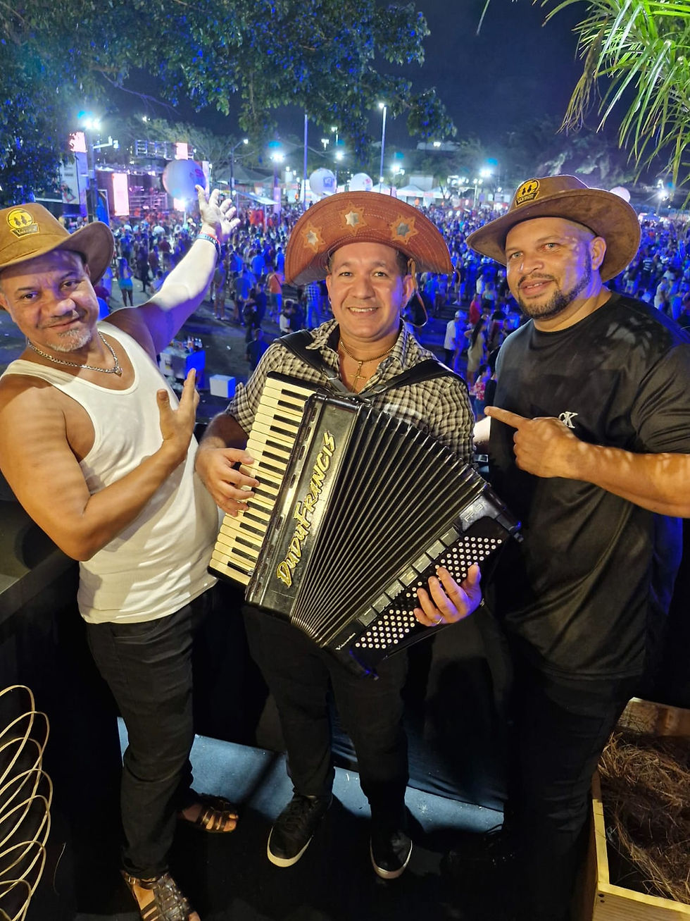 Três homens com chapéus de cangaceiro e caubói posam para foto. Um segura sanfona. Festa noturna ao fundo com muitas luzes.