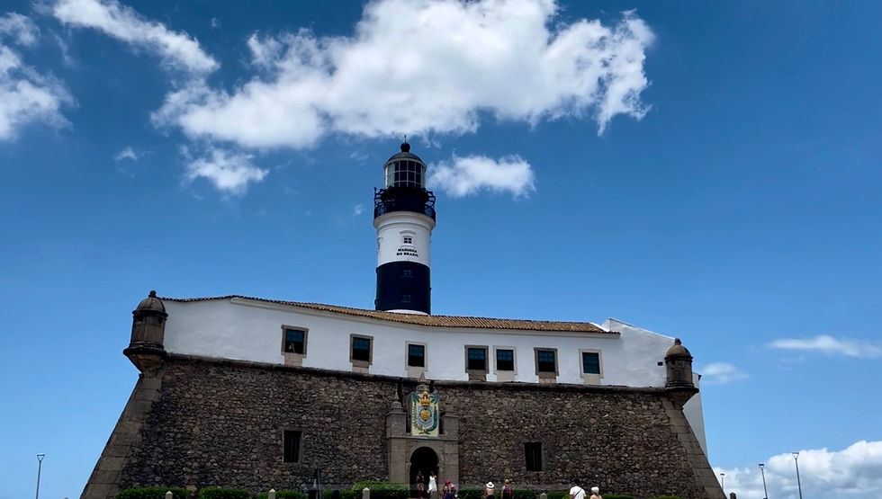 Farol da Barra em Salvador sob céu azul com nuvens. Pessoas visitam o forte histórico de pedra e branco; ambiente sereno e turístico.