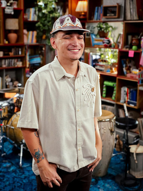 Homem sorrindo, usando chapéu de couro estilizado e camisa clara, em ambiente interno com instrumentos musicais e estantes ao fundo.