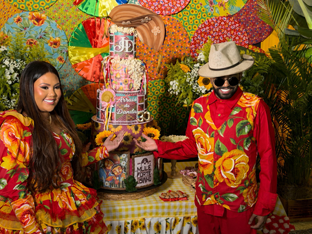 Casal (Jéssica Dandara e Silvanno Salles) sorridente em trajes florais de festa junina posa diante de bolo colorido com texto Arraiá da Dandara.