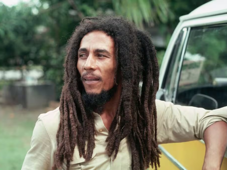 Homem (cantor Bob Marley) com cabelo dreadlock e barba encostado em uma van amarela, olhando pensativo para a direita. Fundo verde e arborizado.