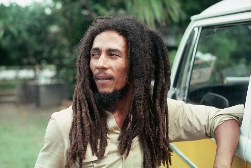 Homem (cantor Bob Marley) com cabelo dreadlock e barba encostado em uma van amarela, olhando pensativo para a direita. Fundo verde e arborizado.