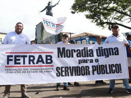 Homens seguram faixa da FETRAB pedindo moradia digna para servidores. Fundo com árvores e edifícios. Atmosfera de protesto. Grupo de servidores públicos segura faixa da Fetrab pedindo moradia digna durante manifestação em Salvador