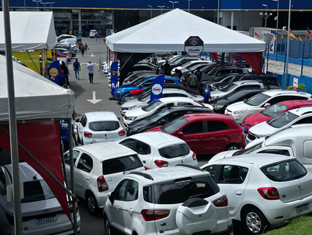 Muitos carros usados sob tendas brancas, com pessoas circulando e placas da Eladio Seminovos em um pátio movimentado.