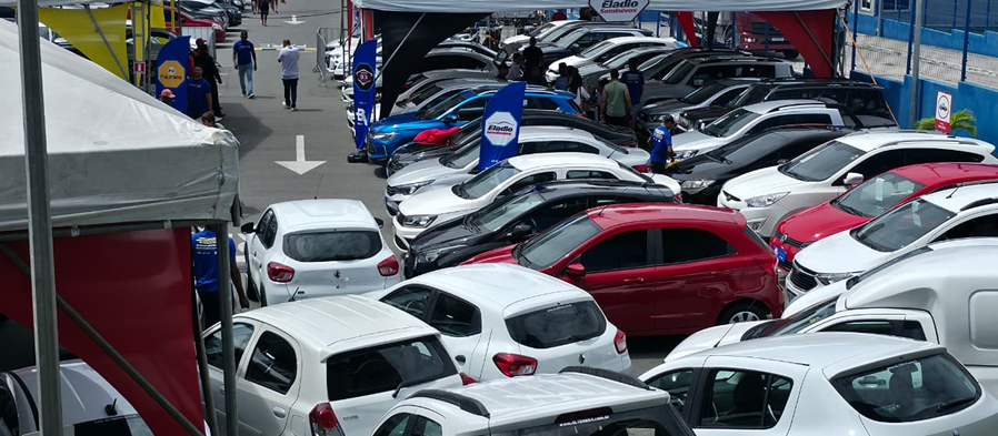 Muitos carros usados sob tendas brancas, com pessoas circulando e placas da Eladio Seminovos em um pátio movimentado.