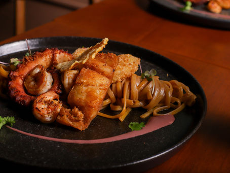 Prato gourmet com polvo grelhado, camarões, massa e crocante sobre prato preto, em mesa de madeira, com decoração delicada