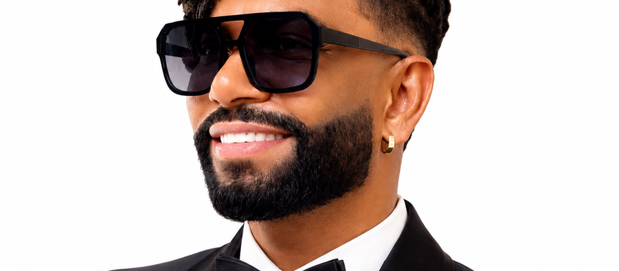 Cantor Silvanno Salles está sorridente de smoking preto e óculos escuros, com dreadlocks e detalhes dourados, em fundo branco.