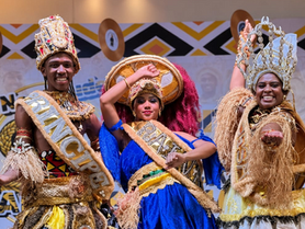 Três pessoas em trajes tradicionais e coroas, sorrindo e posando em palco decorado; faixas douradas com texto visível.