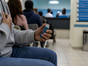 Pessoa segurando inalador azul em sala de espera de hospital. Outras pessoas sentadas ao fundo. Ambiente hospitalar e neutro.