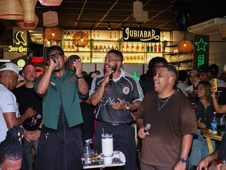 Cantores Lucas e Orelha se apresentam animados com Dan Mocidade em bar lotado, com luzes neon, garrafas ao fundo e placas Jose Cuervo e Jubiabar.