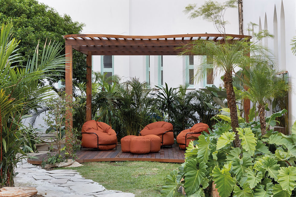 Jardim tropical com pérgola de madeira, poltronas laranja e bancos, cercado por palmeiras e folhagens diante de parede branca.