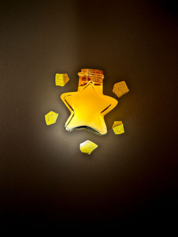 A glowing, star-shaped glass bottle filled with a golden yellow liquid, sealed with a cork. Surrounding the bottle are small, translucent yellow shards that resemble pieces of a magical or crystallized material. The bottle appears to emit a soft light, casting a gentle glow on a dark background.