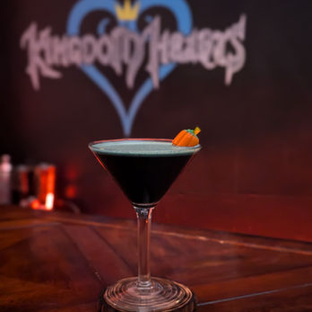 A dark black drink with a white layer on top stands in a martini glass with a small pumpkin-shaped piece of candy corn peeking out from the drink's surface.